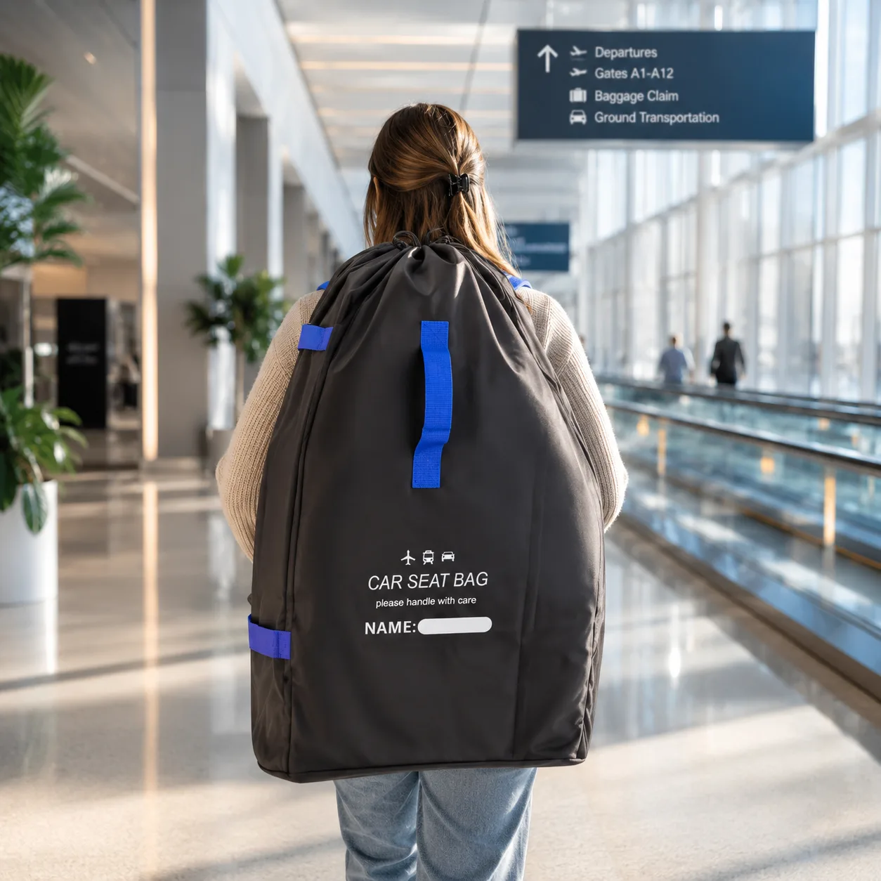 Woman walking through an airport terminal wearing the black car seat travel bag with blue straps and blue side handles, the printed name window visible on the back panel