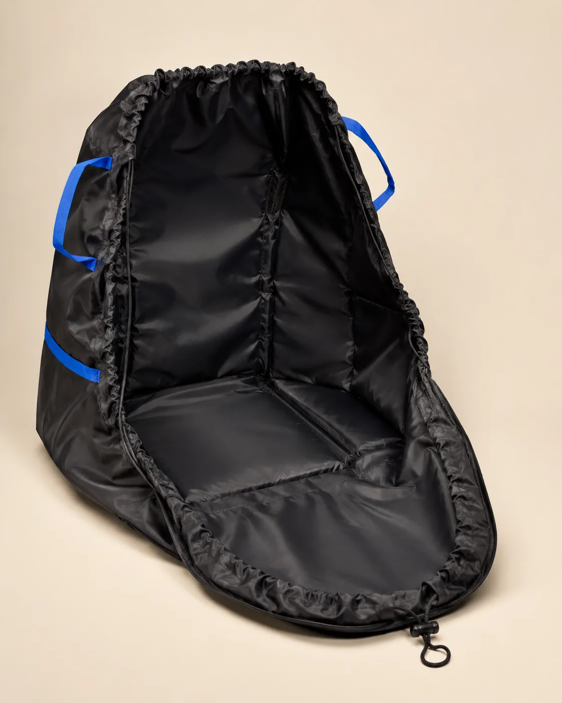 Black car seat travel bag unzipped and laid open flat on a cream studio background, showing the full lined interior, the blue webbing handles and the drawstring closure with its toggle